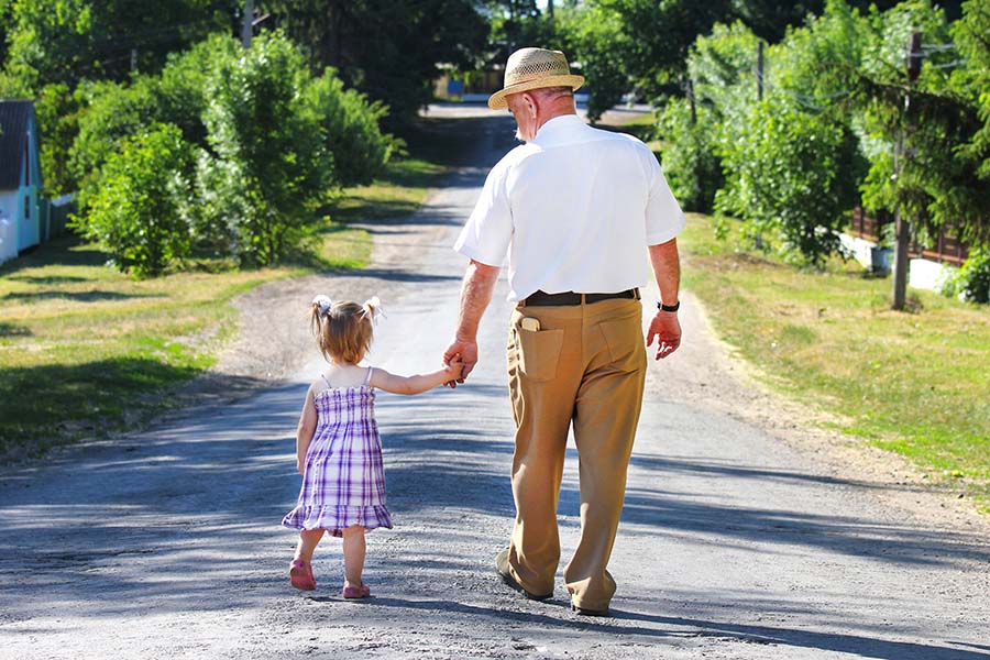 About Our Agency - View of a Grandfather Walking with His Granddaughter on an Empty Country Road While Holding Her Hand